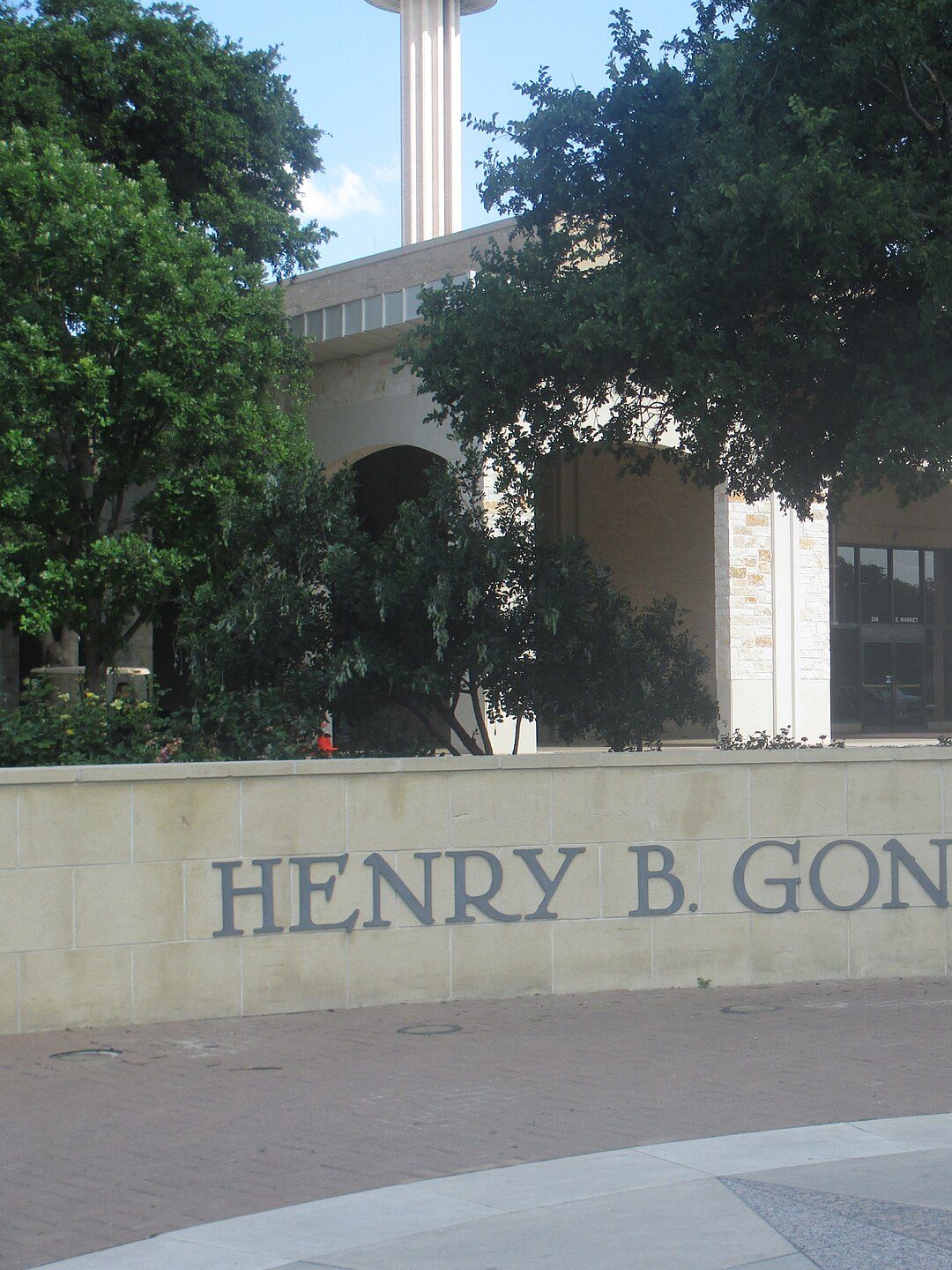 A stone wall lettered Henry B. Gonzalez Convention Center in front of an arched pale stone building, with the column of the Tower of the Americas rising behind it