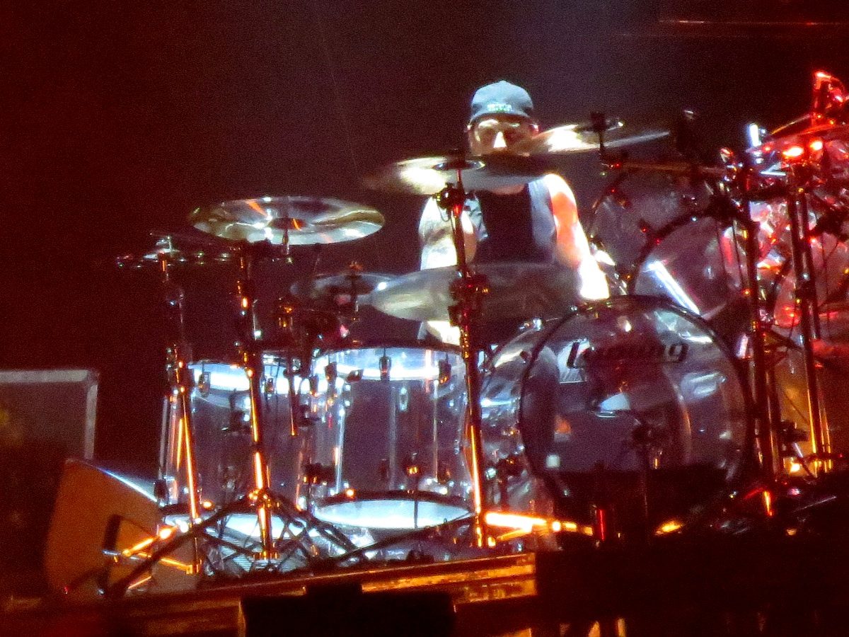 Dave Lombardo in a cap and sleeveless shirt behind a clear acrylic Ludwig drum kit lit orange and white on a dark stage