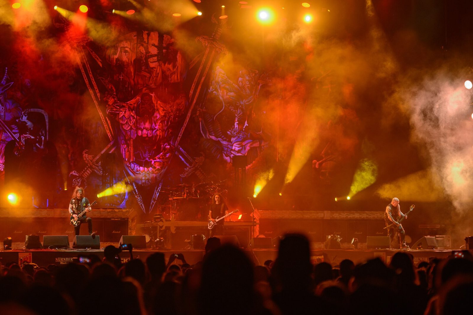 Slayer on a festival stage at night, three players spread across the front under orange and red lights, a giant skull-and-crossed-swords backdrop behind the drum kit and the crowd in silhouette below