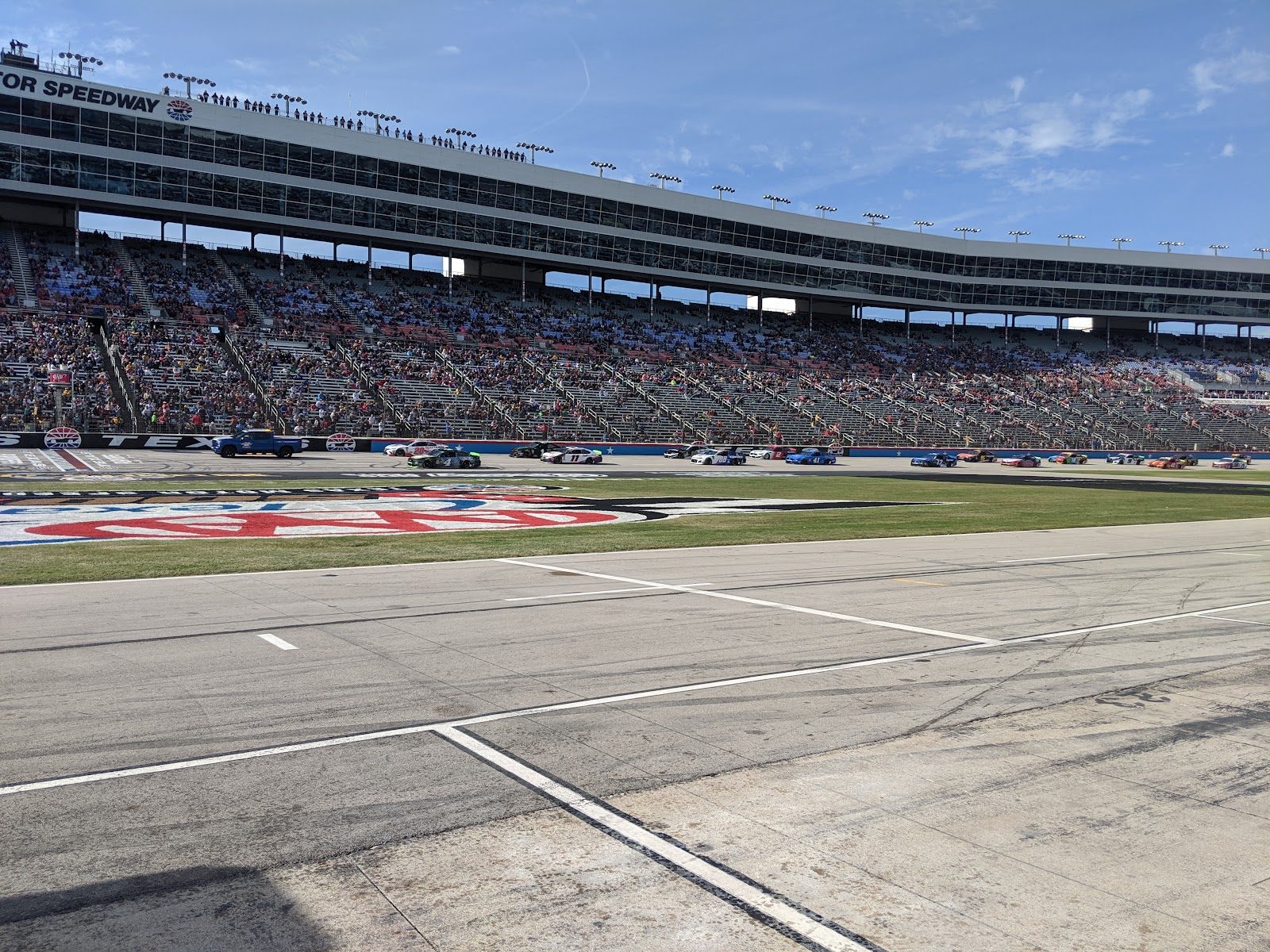 Texas Motor Speedway seen from pit road during a NASCAR race, stock cars on the track in front of a long, packed grandstand under a blue sky