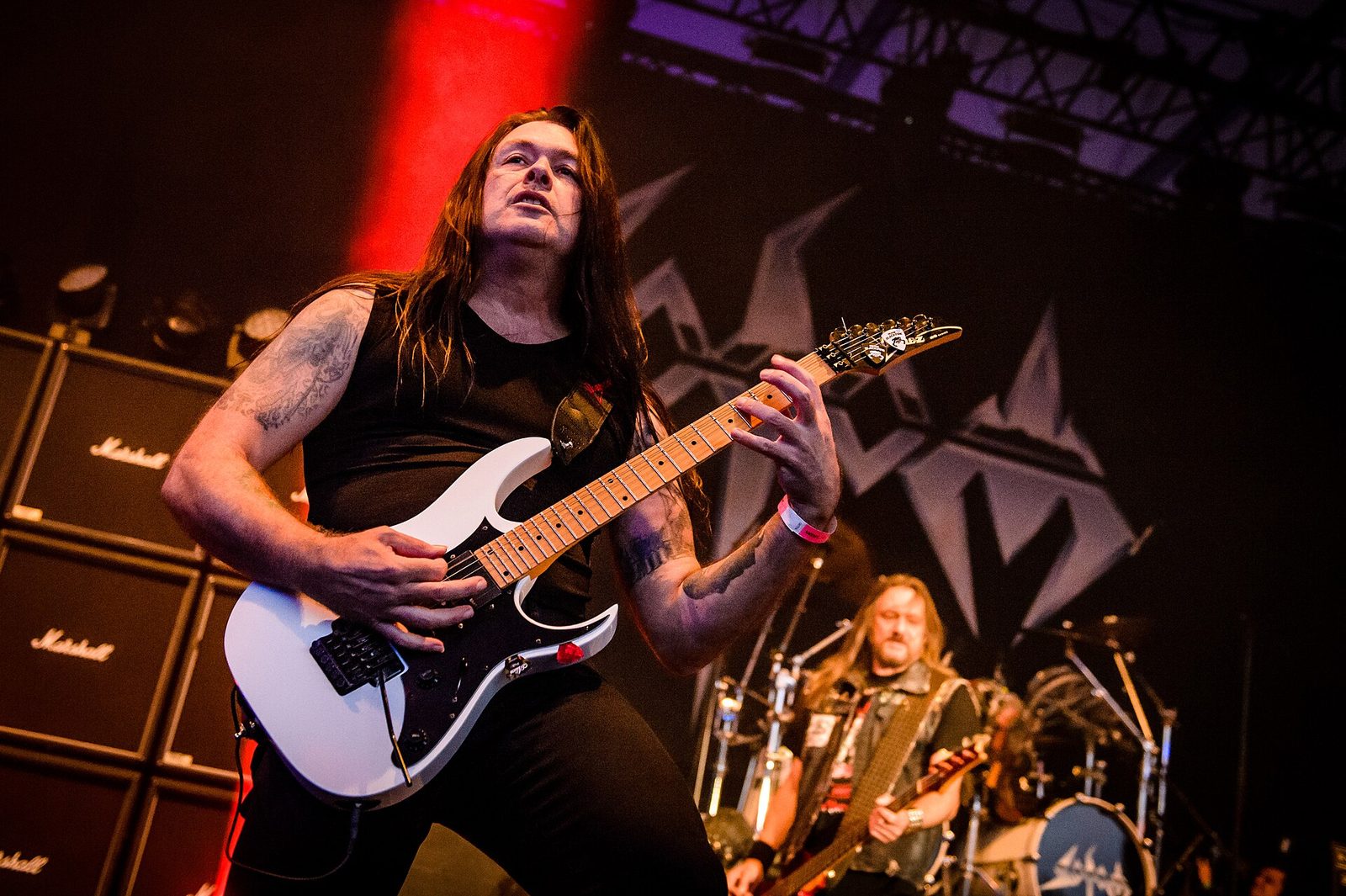 Frank Blackfire, long dark hair and a sleeveless black shirt, playing a white guitar in front of Marshall cabinets and the Sodom logo, bandmates behind him