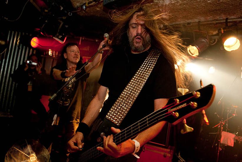 Sodom on a club stage under red light, a long-haired bassist in a studded strap in the foreground and a guitarist behind him