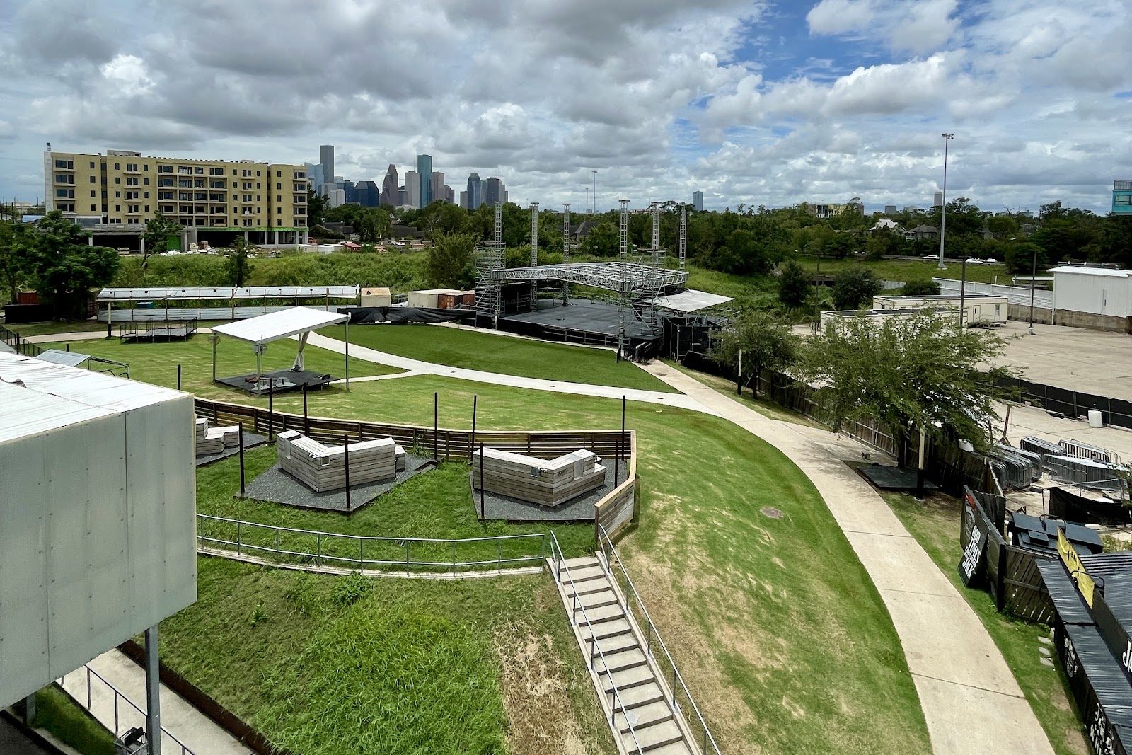 White Oak Music Hall's outdoor Lawn by day, seen from above: terraced grass and a walkway leading to the empty steel-truss stage, with the downtown Houston skyline behind