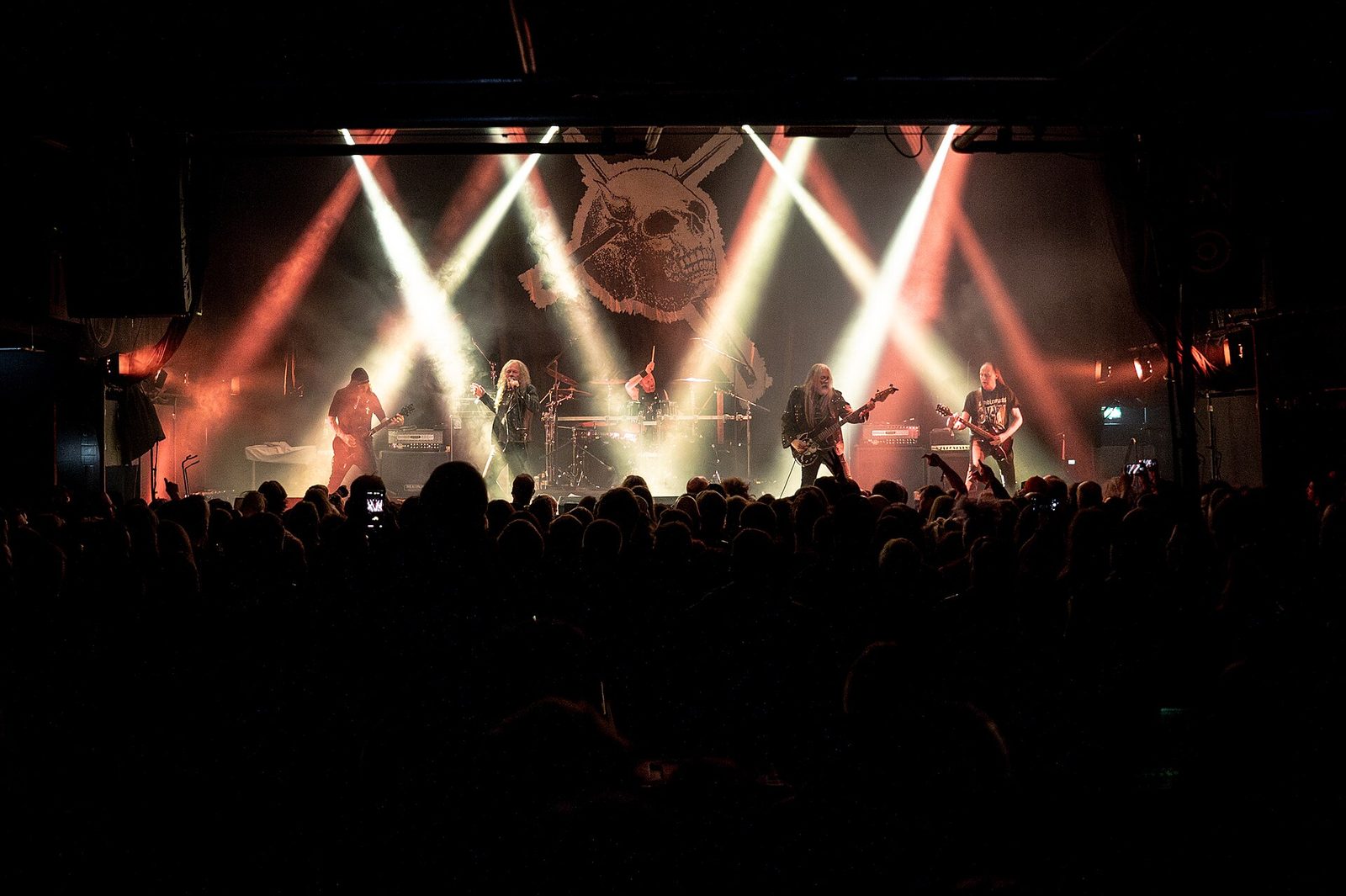 Candlemass performing on a smoky stage under crossing white and red spotlights, a large skull banner behind the five members and a packed crowd in silhouette