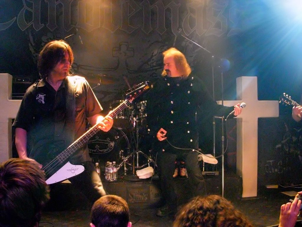 Robert Lowe singing into a hand-held microphone beside bassist Leif Edling on a Candlemass stage with white crosses and the Candlemass logo behind them, 2008