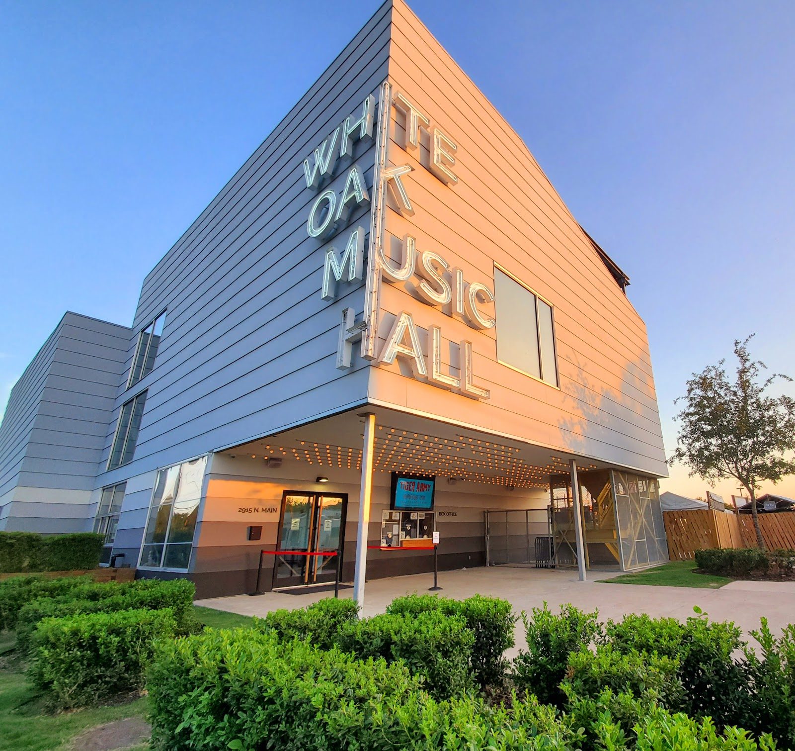 The silver-paneled White Oak Music Hall building in Houston, its name in large white letters on the corner above the glass entrance, with hedges in front