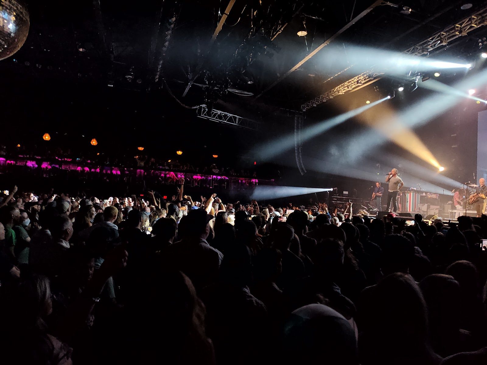A packed main floor at the Bomb Factory seen from the crowd, white spotlights sweeping over a band on the stage at right and a lit balcony along the back wall