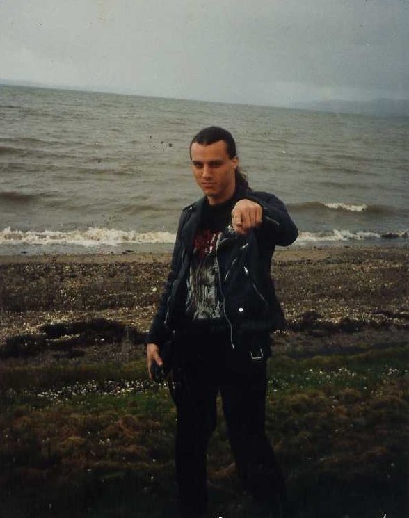 Chuck Schuldiner in a black leather jacket and band T-shirt, pointing at the camera on a pebbly shoreline with gray sea behind him