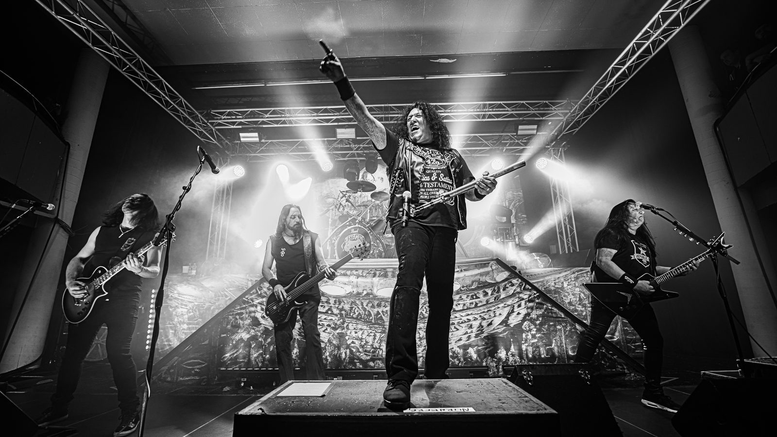 Black and white photo of Testament on a club stage: Chuck Billy in the center on a riser pointing upward, with two guitarists and a bassist spread across the stage under bright stage lights