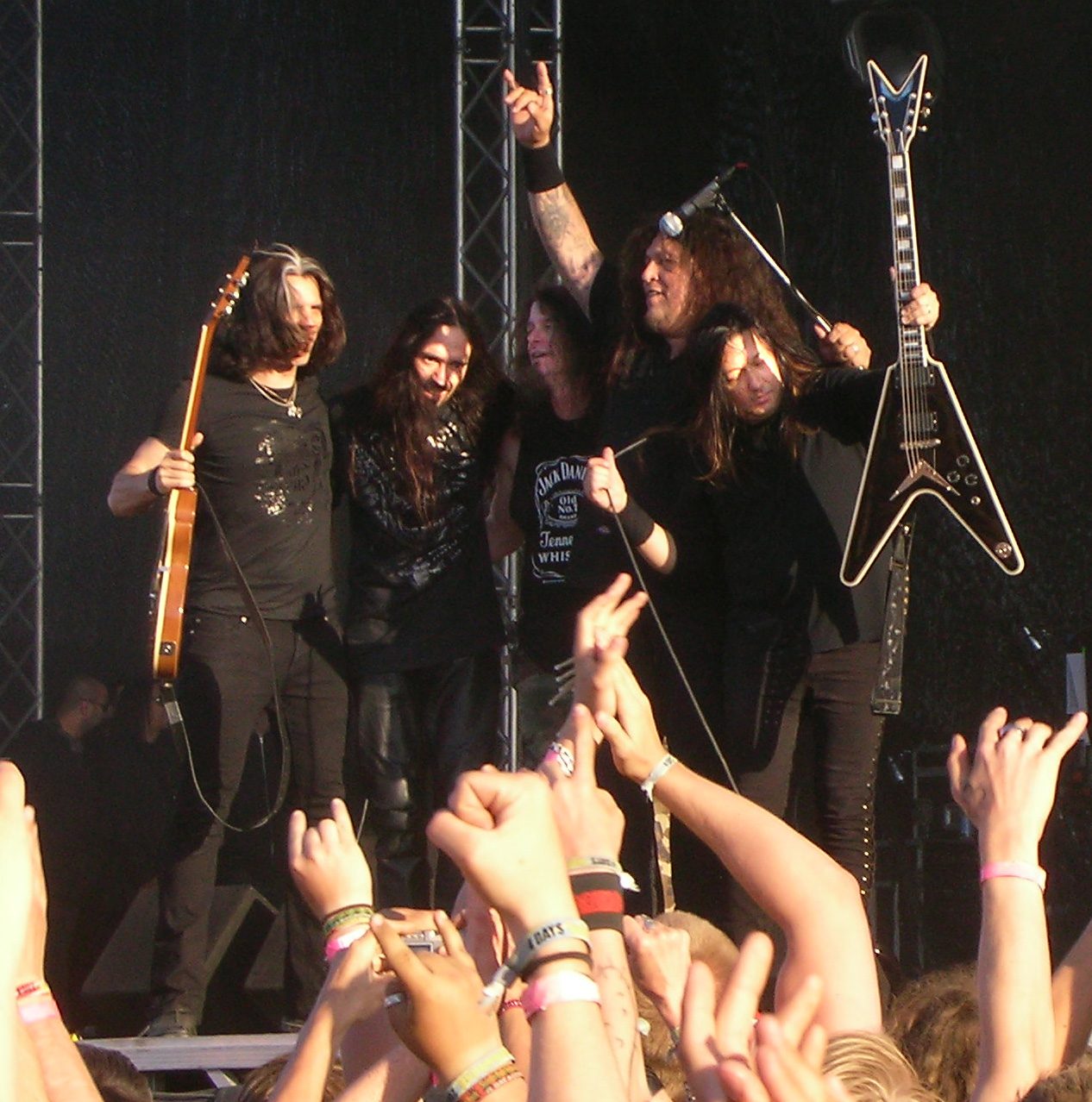Testament lined up at the front of an outdoor festival stage, one member holding a natural-finish Les Paul, another a black V-shaped guitar with white edging, Chuck Billy raising a fist, with fans' hands raised in the foreground