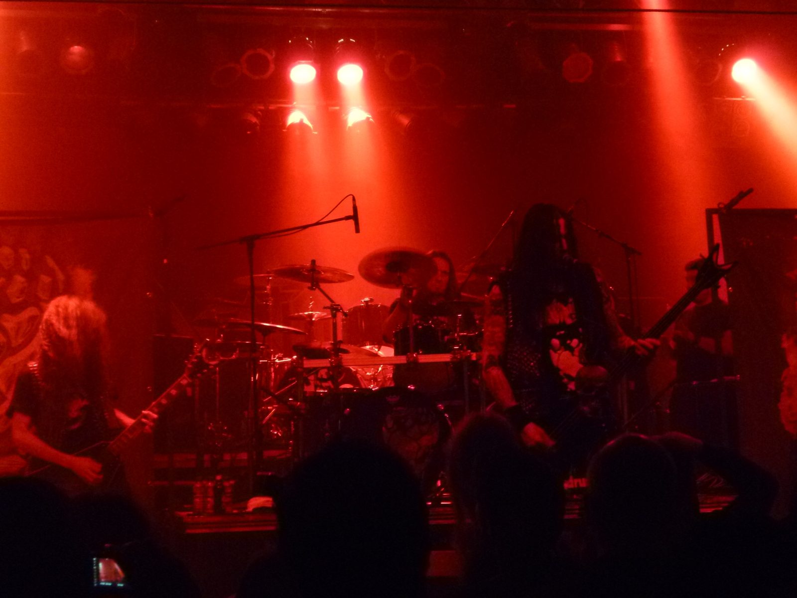 Destruction on a red-lit club stage, a guitarist at left, the drummer behind the kit and Schmier at right with his bass