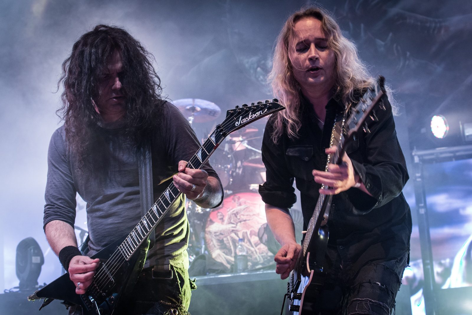 Two Kreator guitarists playing side by side through stage smoke, one dark-haired with a black Jackson V, one blond with long hair, the drum kit behind them
