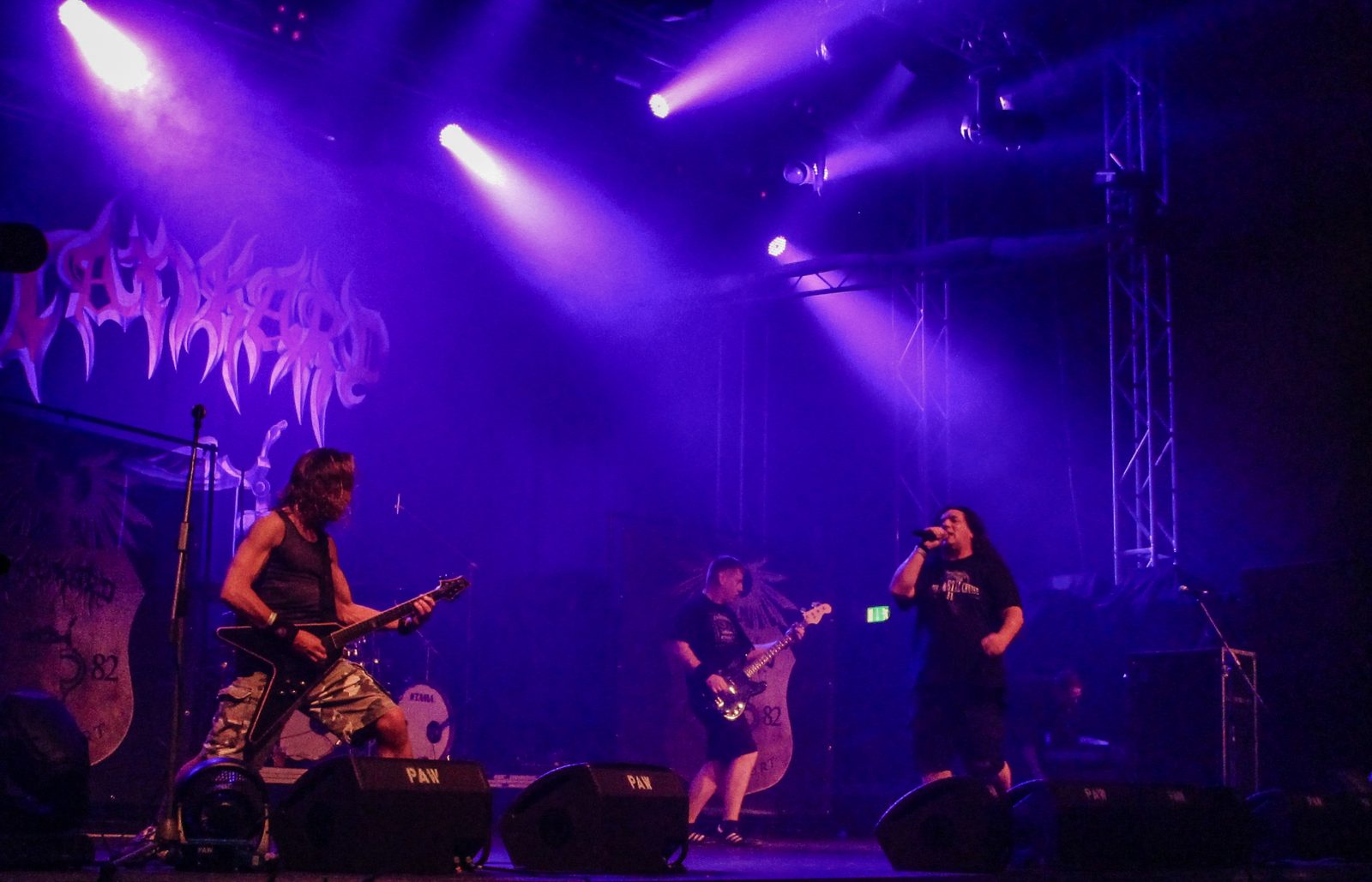 Tankard on a purple-lit stage in front of the band’s logo backdrop, a guitarist with a black Flying V at left, the bassist in the middle and the singer at right with a microphone