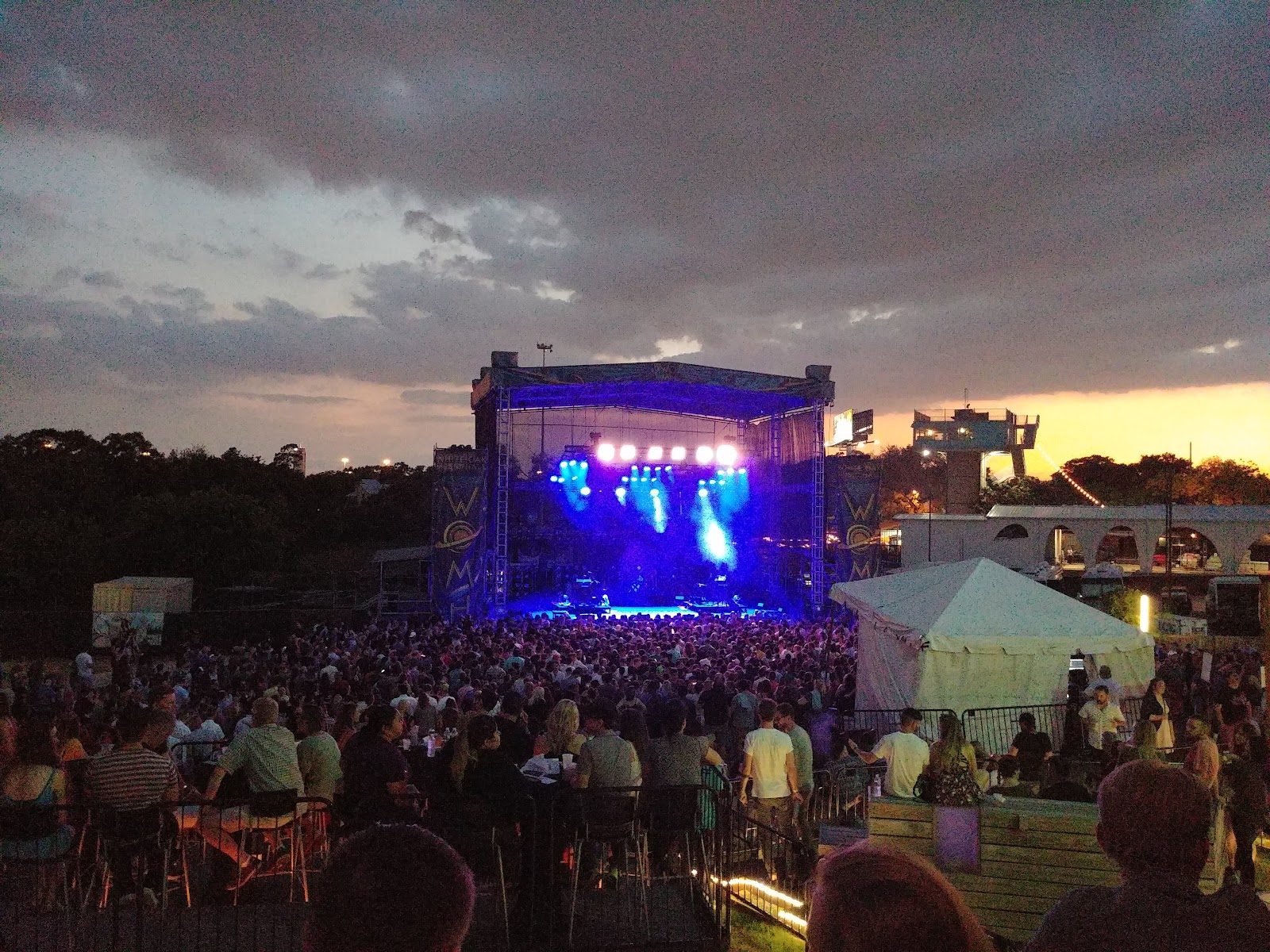 The outdoor lawn stage at White Oak Music Hall at dusk, lit blue and purple, with a large crowd in front and storm clouds overhead