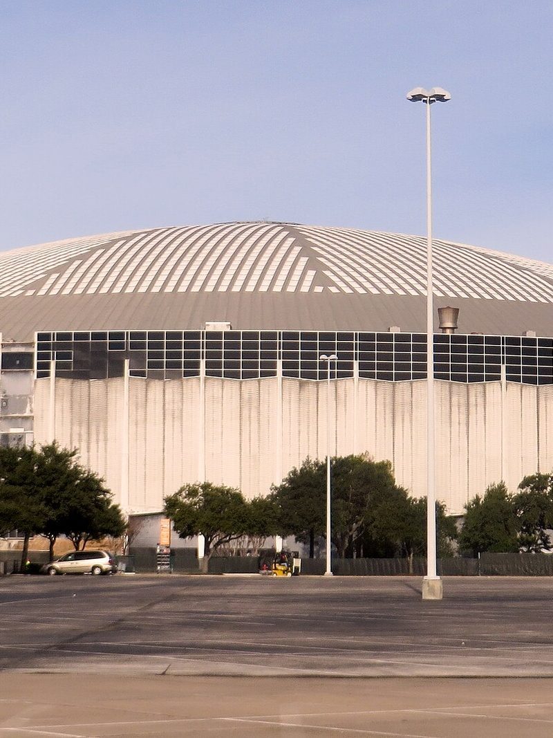 The Houston Astrodome's low white dome and ribbed concrete walls seen across an empty parking lot, with a row of trees along its base