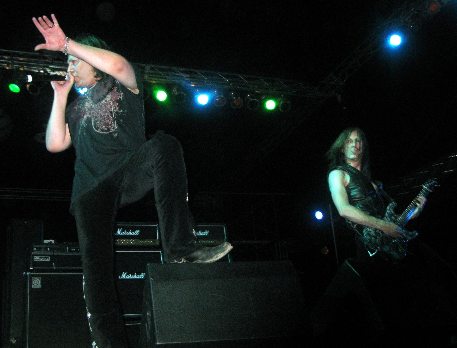 Don Dokken singing with one foot up on a stage monitor while guitarist Jon Levin plays behind him under blue and green stage lights