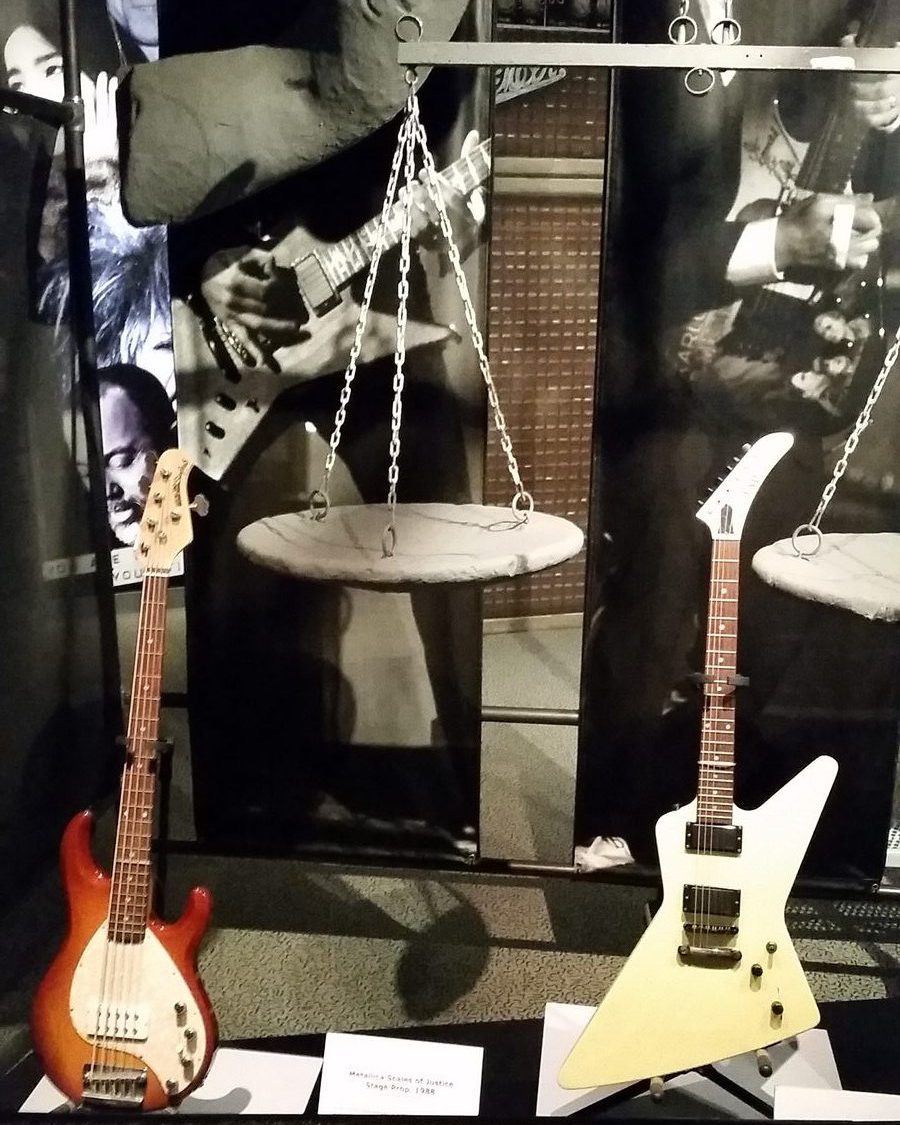 Museum display of a stone-look scales of justice stage prop hanging on chains, with a white ESP Explorer guitar and a sunburst five-string bass on stands in front