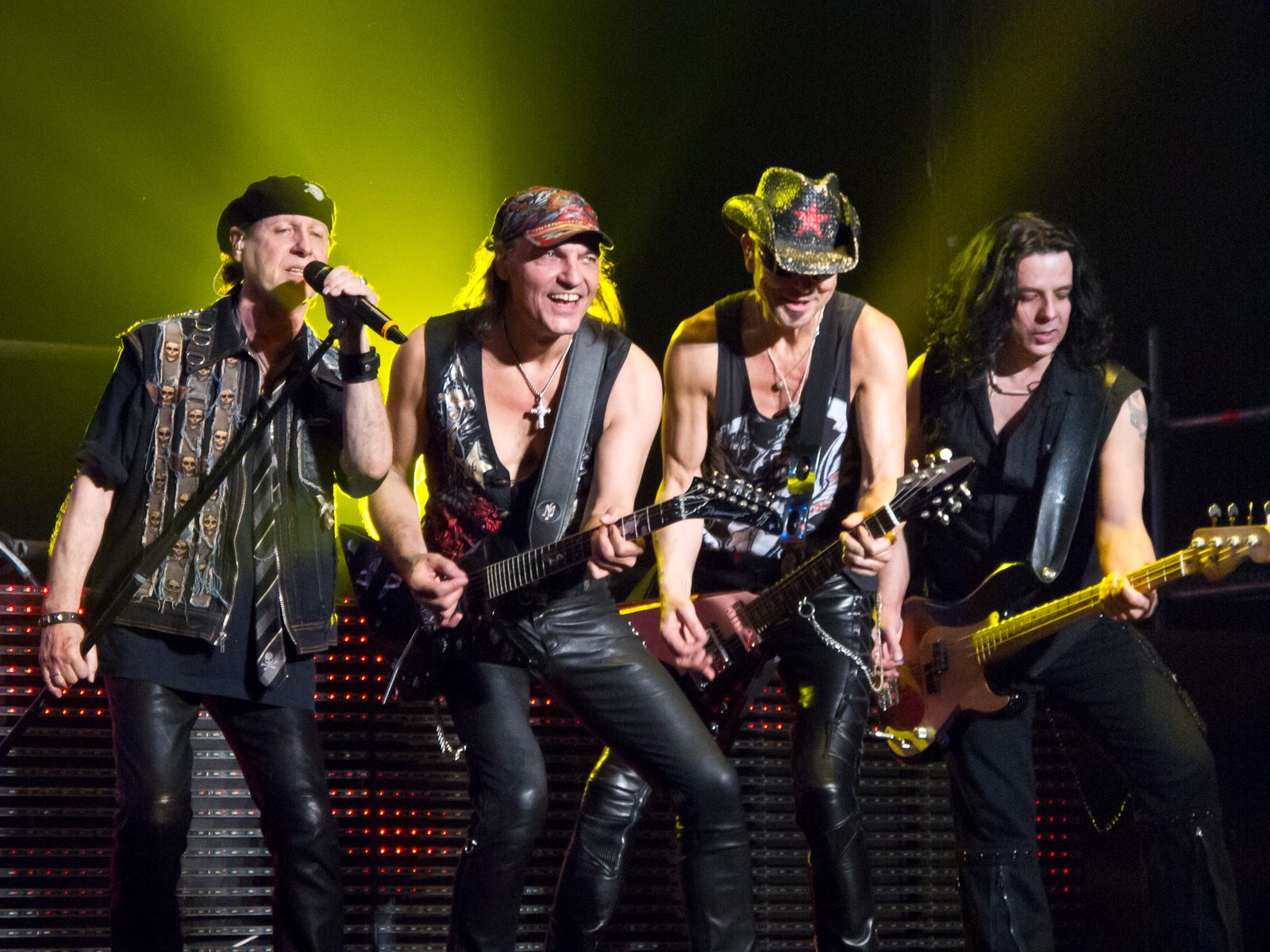 Four members of the Scorpions lined up at the front of the stage under yellow-green light, Klaus Meine singing on the left with three guitarists beside him