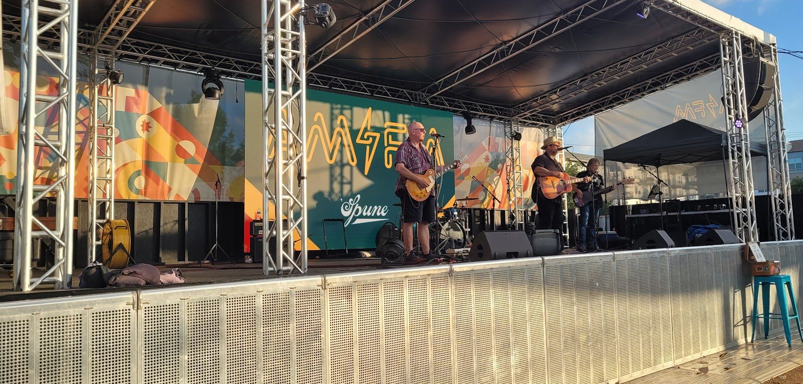 An outdoor covered stage at AM/FM in Dallas with metal truss framing, a band playing guitars and drums, and a large AM/FM sign on the backdrop behind them