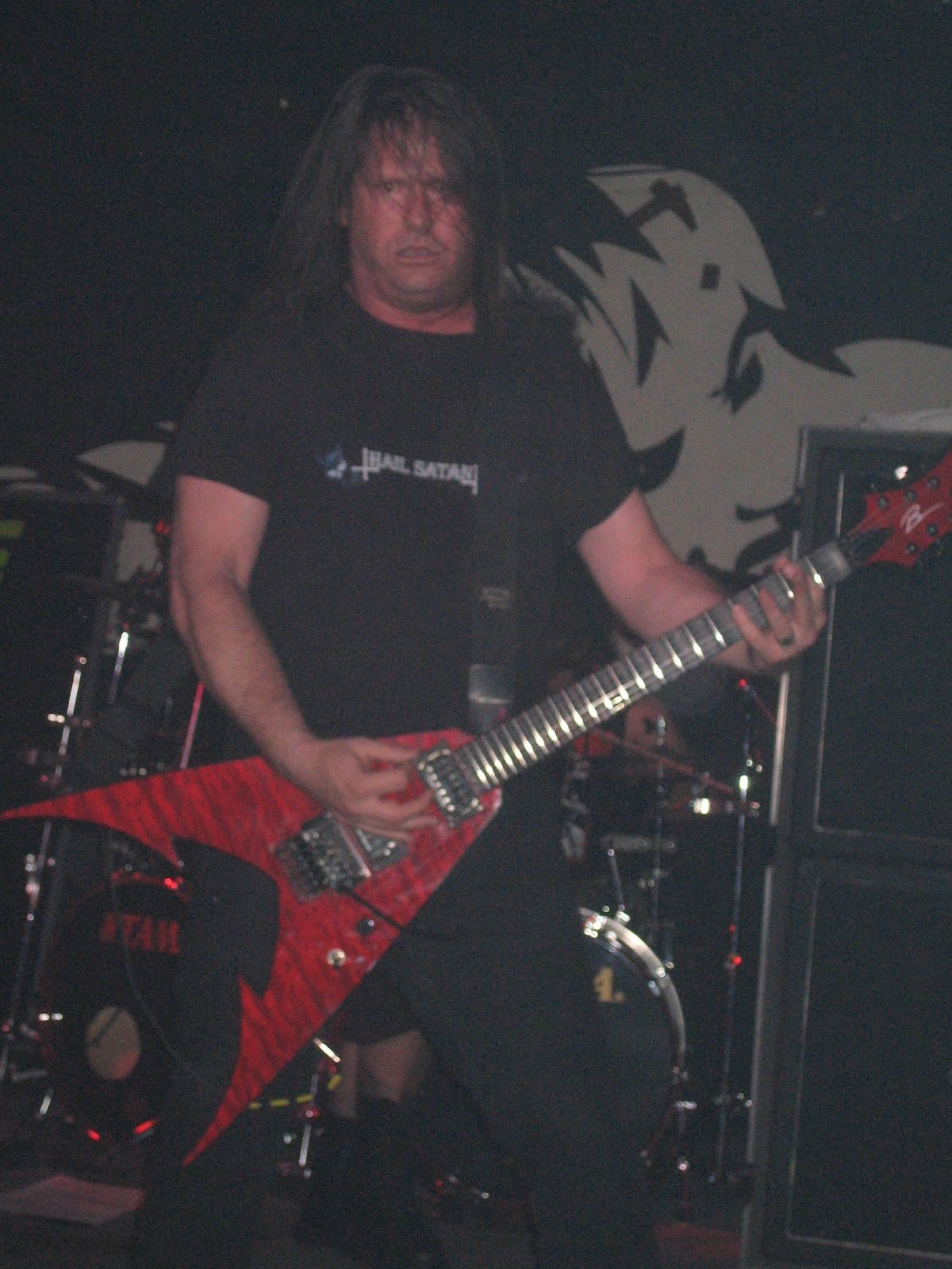 Exodus guitarist Gary Holt in a black T-shirt playing a red flame-shaped V guitar on a dark club stage, drum kit and amplifier stack behind him