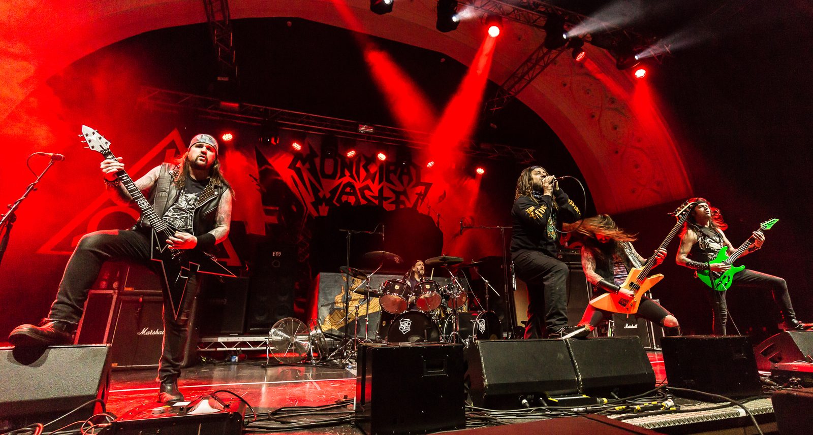 Municipal Waste on a wide stage bathed in red light: the singer in the center with a microphone, four guitarists and bassists spread across the front and the drummer behind, the band's logo on the backdrop