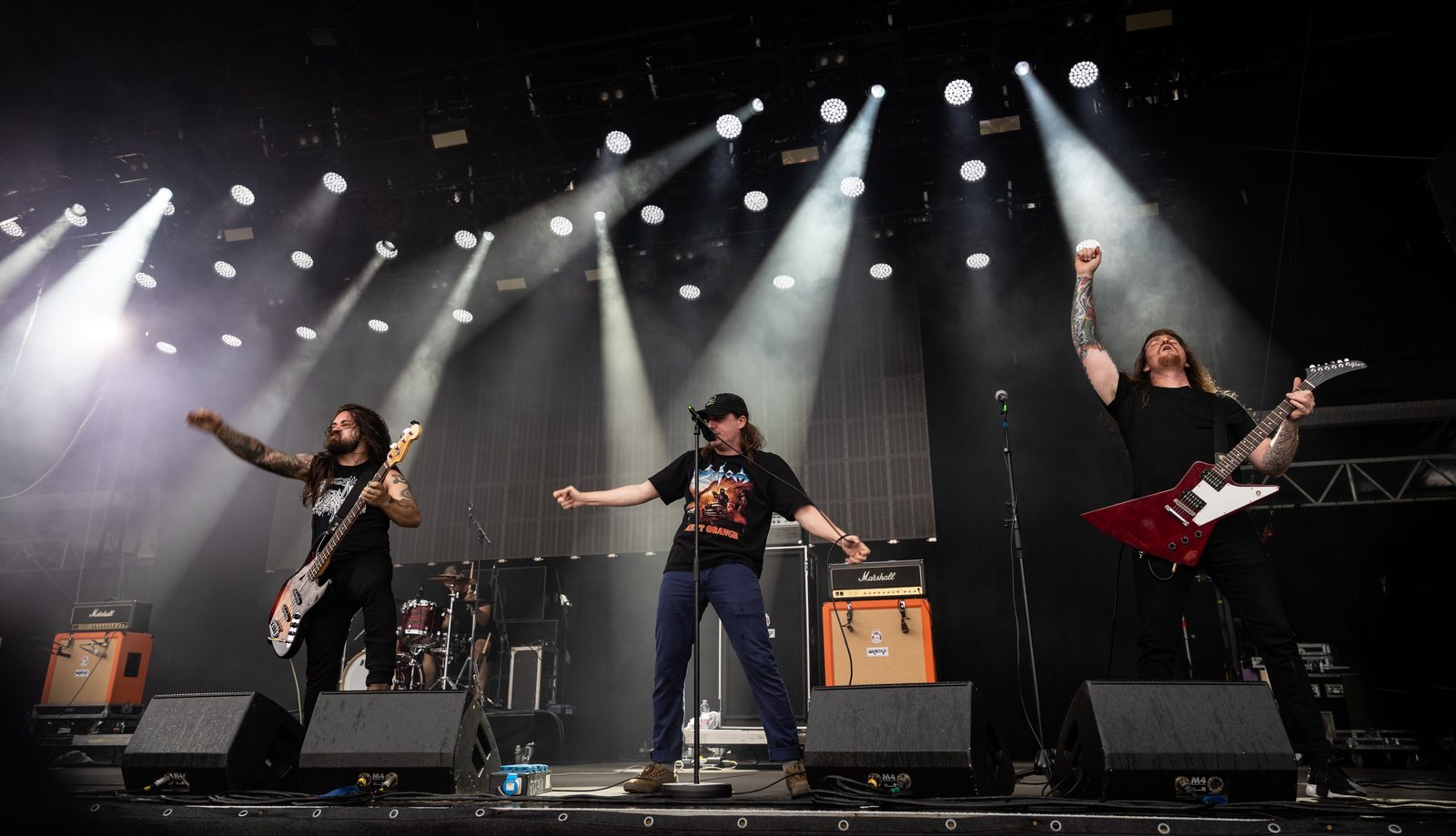 Power Trip's singer in a cap and T-shirt with arms spread at the microphone, a bassist and a guitarist with a red Explorer on either side under white spotlights