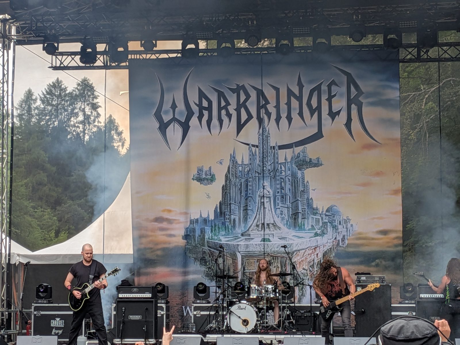 Warbringer on an outdoor festival stage in front of a huge backdrop with the band's logo over a floating castle, pine trees at the side