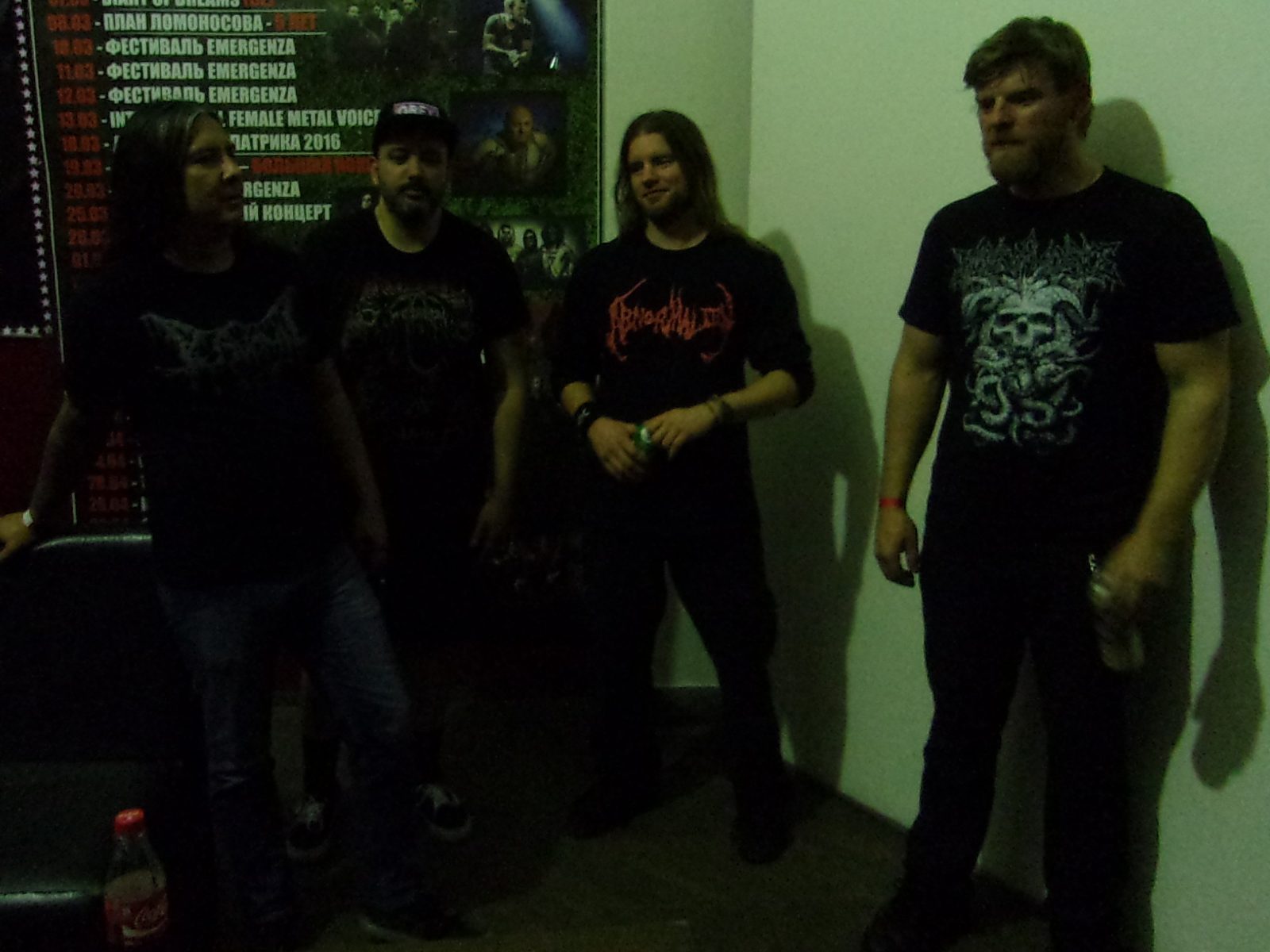 Four members of Devourment in black band T-shirts standing backstage against a white wall beside a Russian-language concert poster