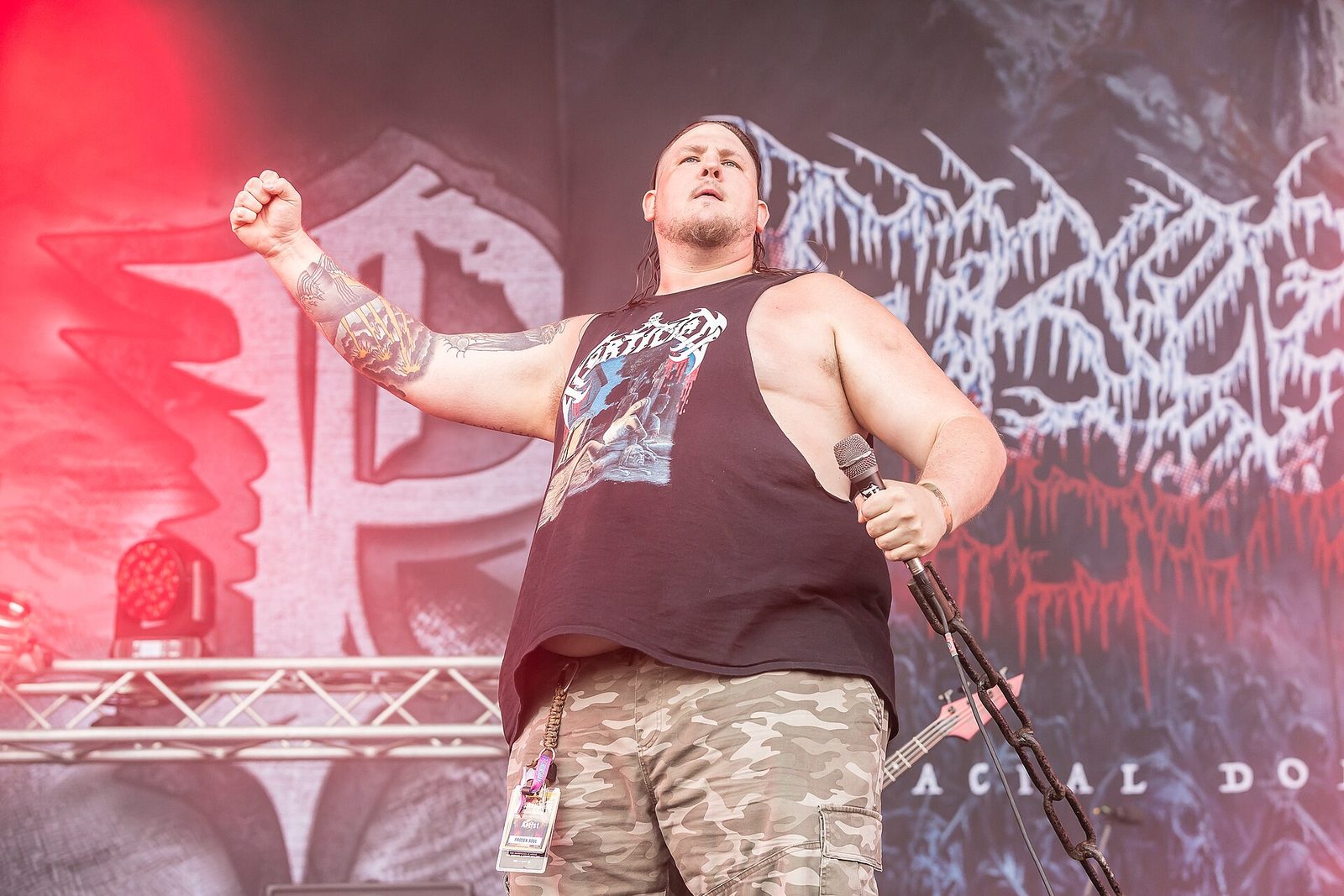 Frozen Soul's singer on an outdoor festival stage, one fist raised, a microphone and a length of chain in the other hand, wearing a sleeveless band shirt and camo shorts in front of the band's icy logo backdrop
