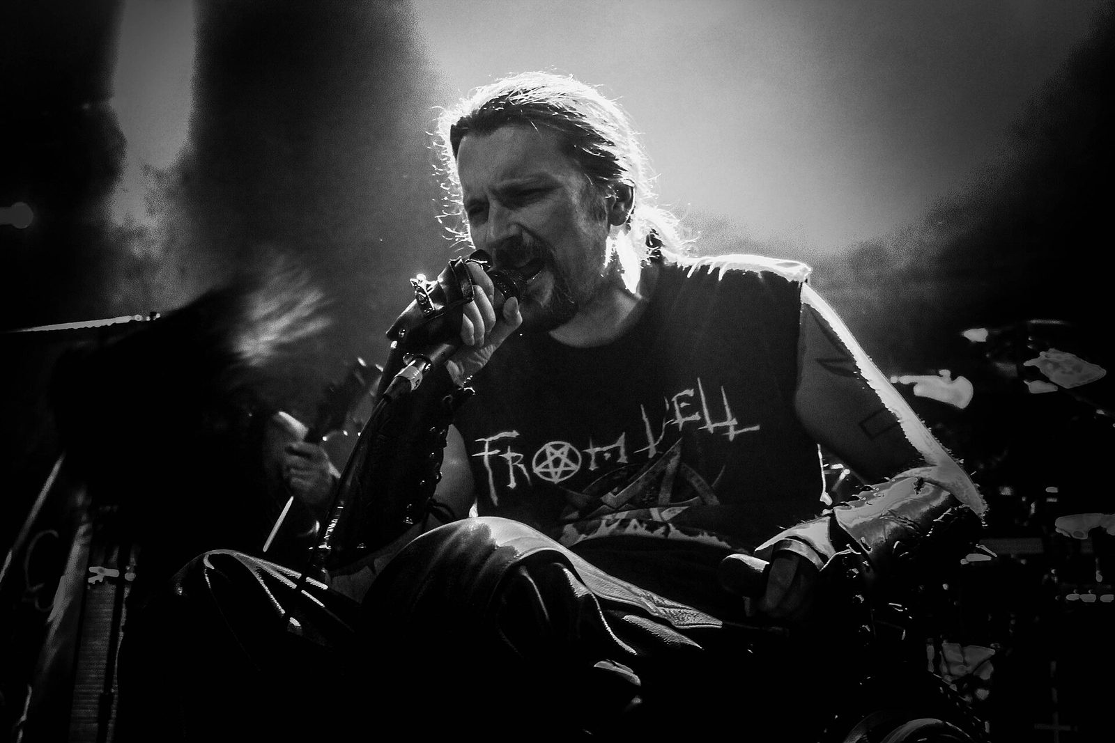 Black-and-white photo of Possessed singer Jeff Becerra on stage, long hair tied back, gripping a microphone in a gauntleted hand and singing, wearing a sleeveless From Hell shirt, with stage smoke and a drum kit behind him
