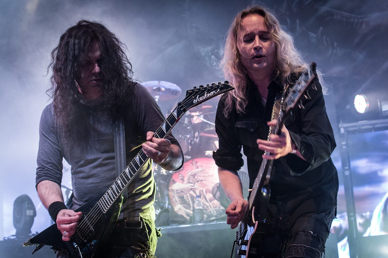 Two Kreator guitarists playing side by side in stage smoke, one with long dark curly hair on a black Jackson V guitar, the other with long blond hair on a spiked black guitar, a drum kit behind them
