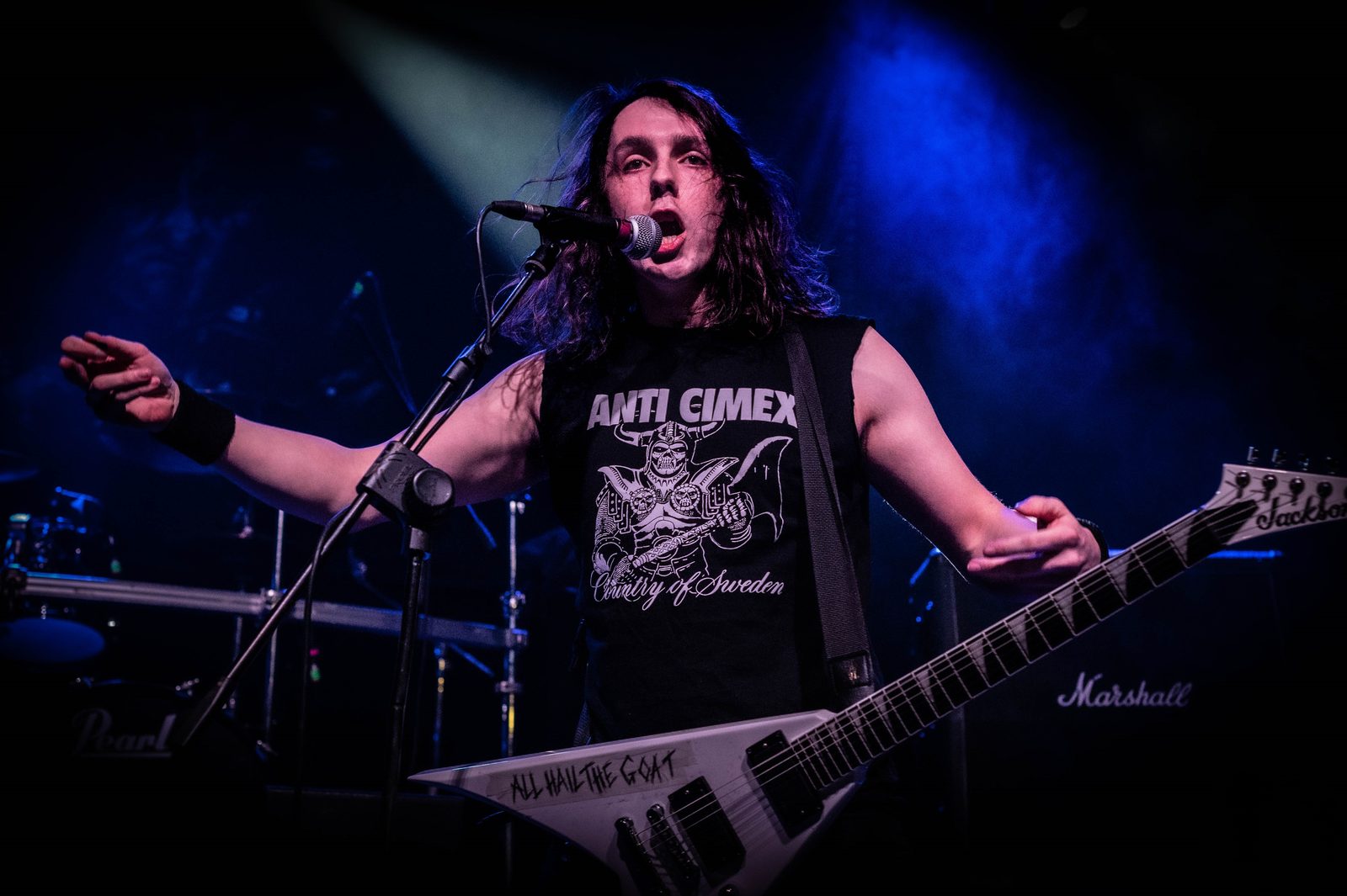 Hellripper's long-haired singer and guitarist singing into a microphone with arms spread, a white V-shaped Jackson guitar marked All Hail the Goat across his body, under blue stage light