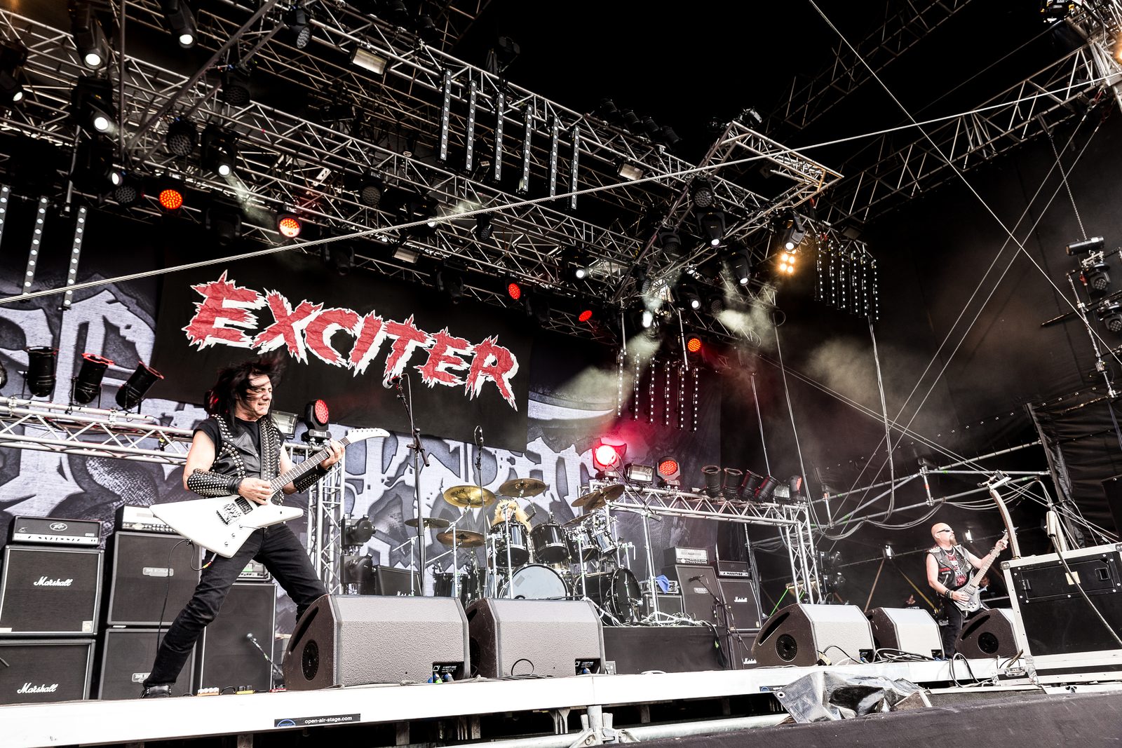 Exciter on an outdoor festival stage under a lighting rig, a long-haired guitarist in a studded vest playing a white Explorer-style guitar on the left, a drum kit in the middle and a bald bassist on the right, with a red and white Exciter banner behind them