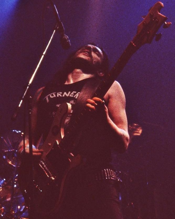 Lemmy of Motörhead on a purple-lit stage, head tipped back toward the microphone, playing a bass guitar in a black sleeveless shirt and studded belt