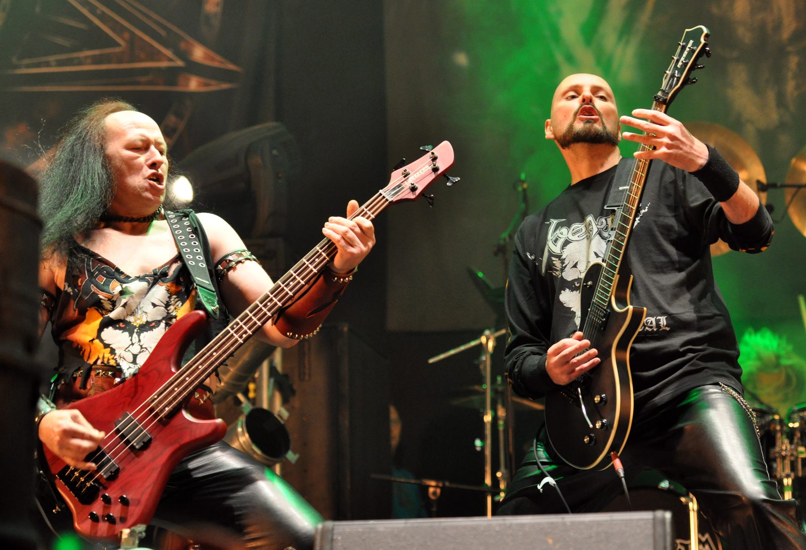 Venom's bassist in a ripped tank top playing a red bass and its guitarist in a black long-sleeved shirt playing a black guitar, under green stage light