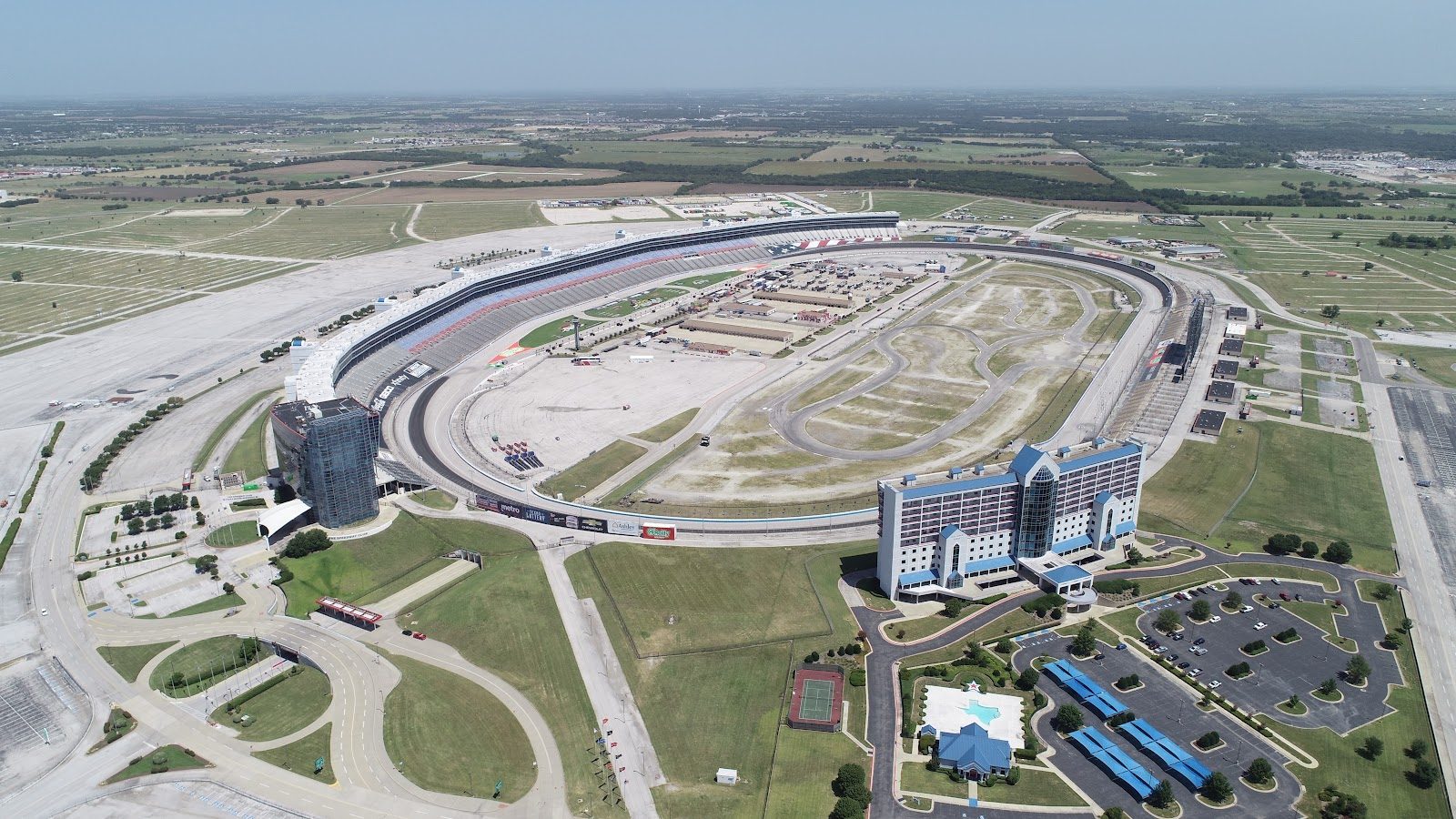 Aerial view of Texas Motor Speedway, an oval track with a long grandstand on one side, a glass tower and a blue-roofed hotel in the foreground, and flat North Texas farmland to the horizon