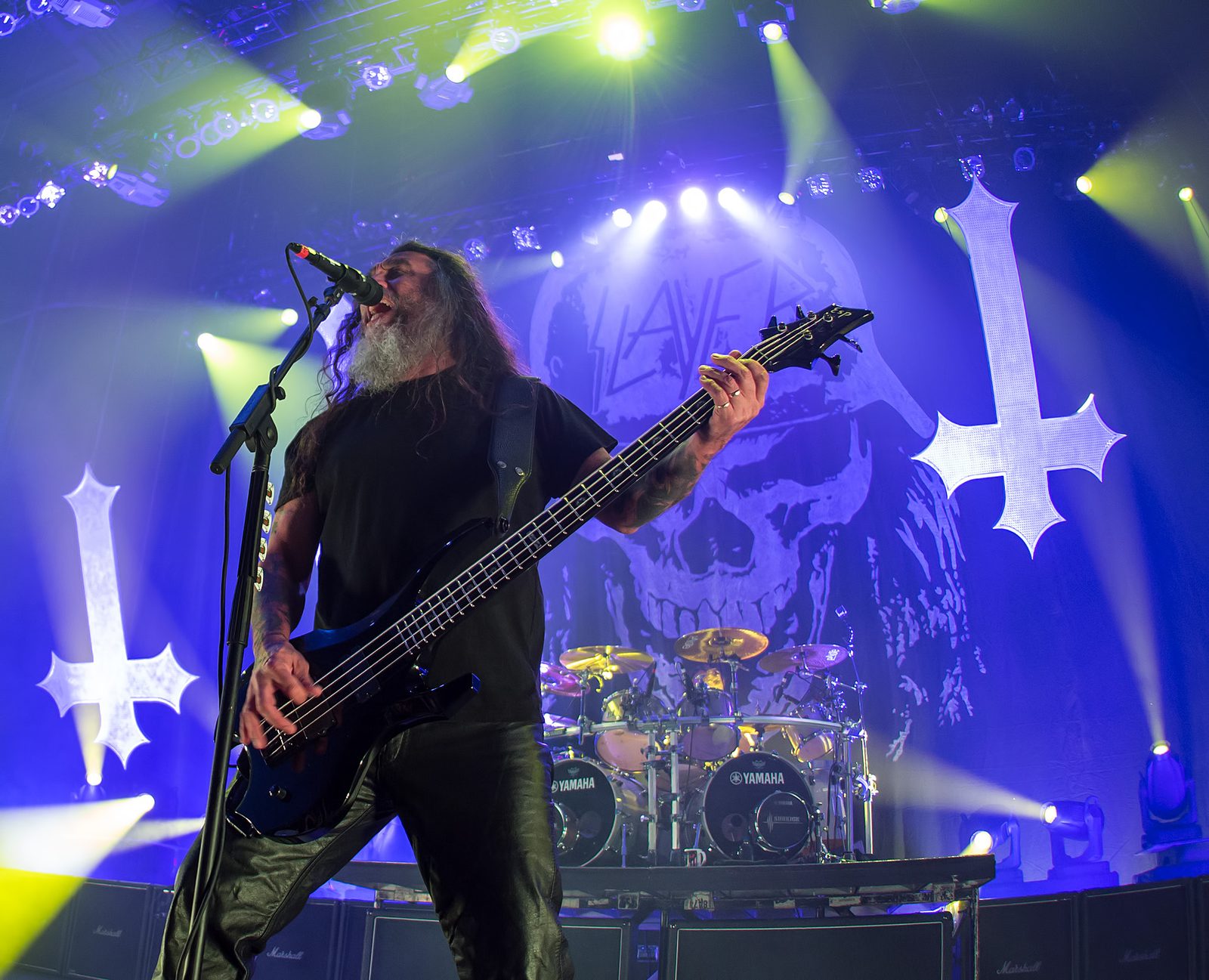 Tom Araya, with long dark hair and a grey beard, singing into a microphone while playing a black bass under blue stage lights, with a Slayer skull backdrop, two white inverted crosses and a Yamaha drum kit behind him
