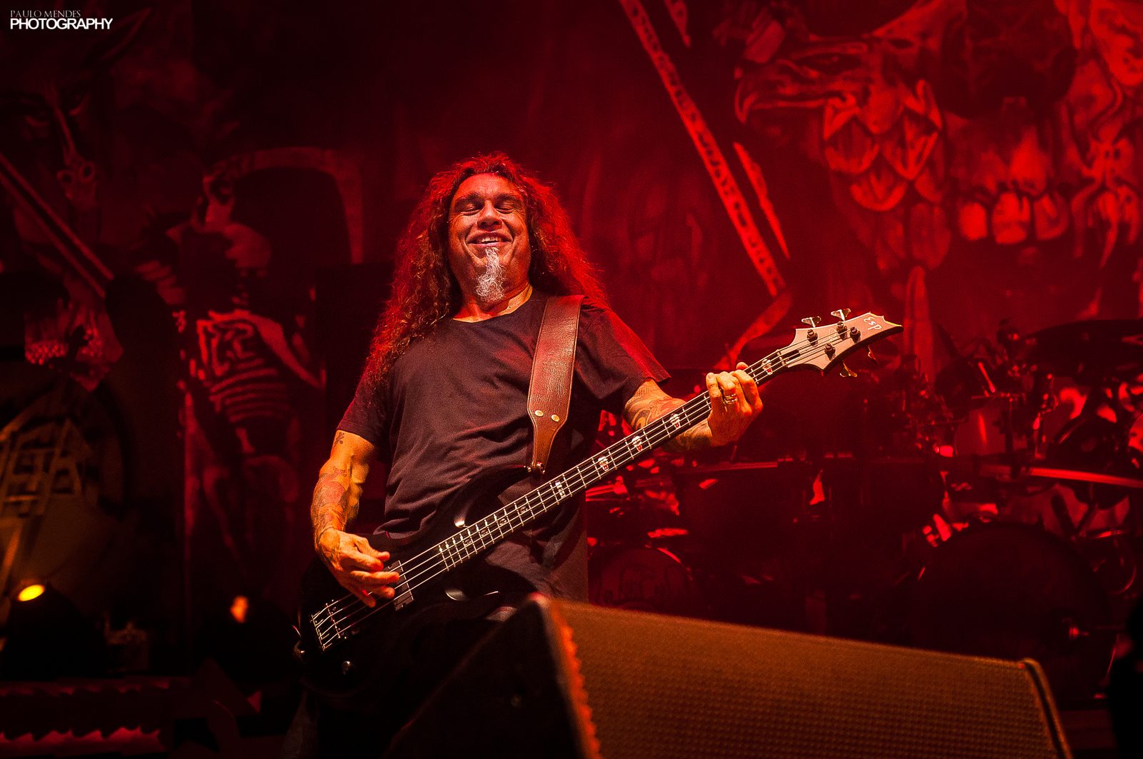 Tom Araya smiling with his eyes closed, playing a black bass with an ESP logo on the headstock, under red stage light with a painted eagle backdrop behind him