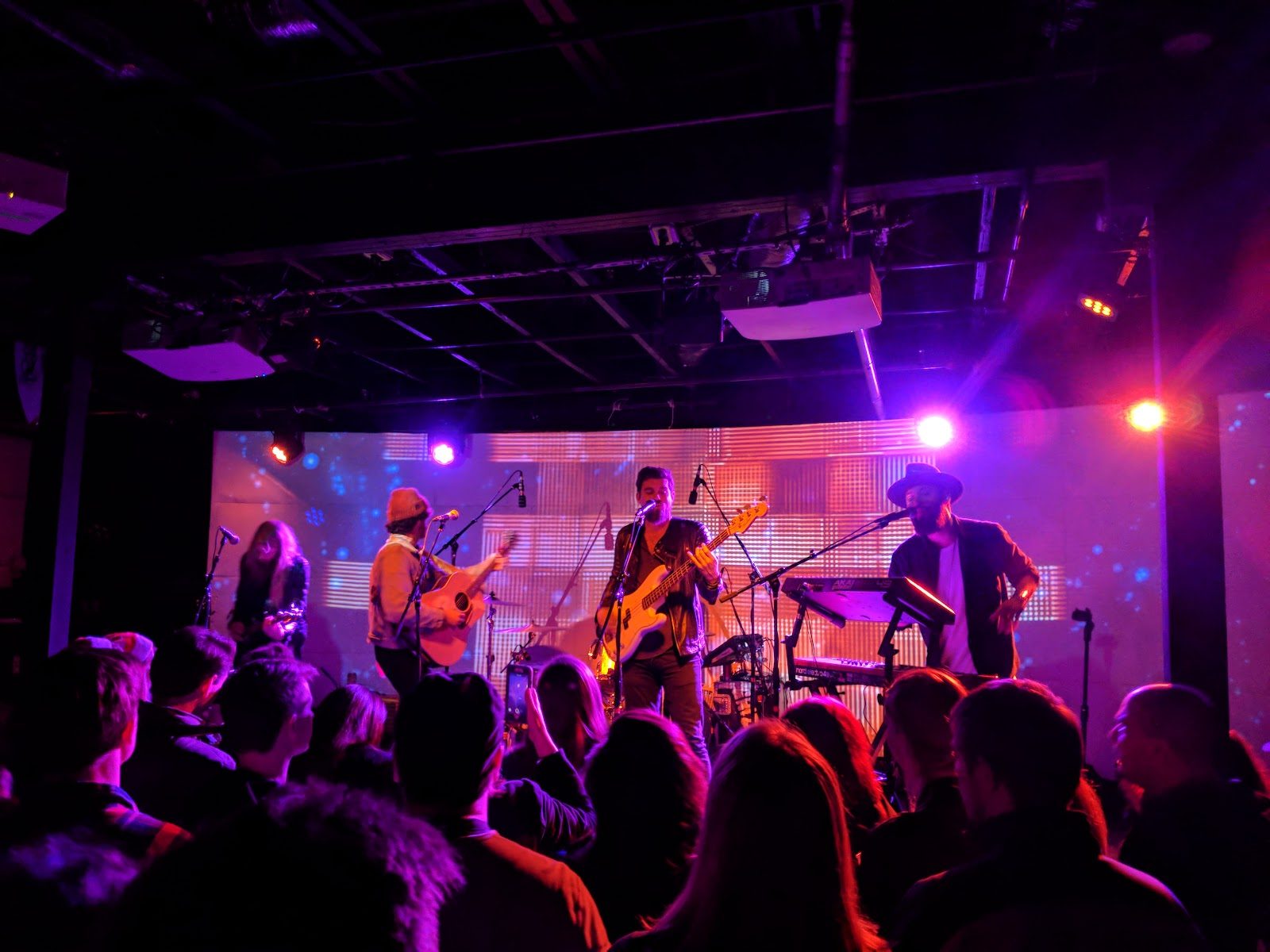 A five-piece band playing on Empire's low stage in Austin under purple and orange lights, with a projected screen behind them and the crowd's heads in the foreground