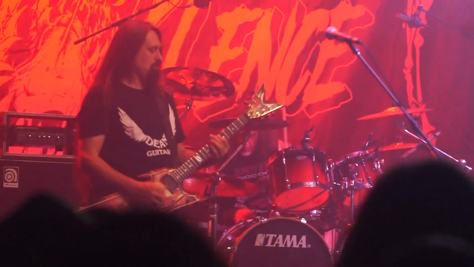 A long-haired guitarist in a Dean Guitars T-shirt playing a gold-topped guitar in front of a red Vio-lence backdrop, with the drummer behind his kit in the background