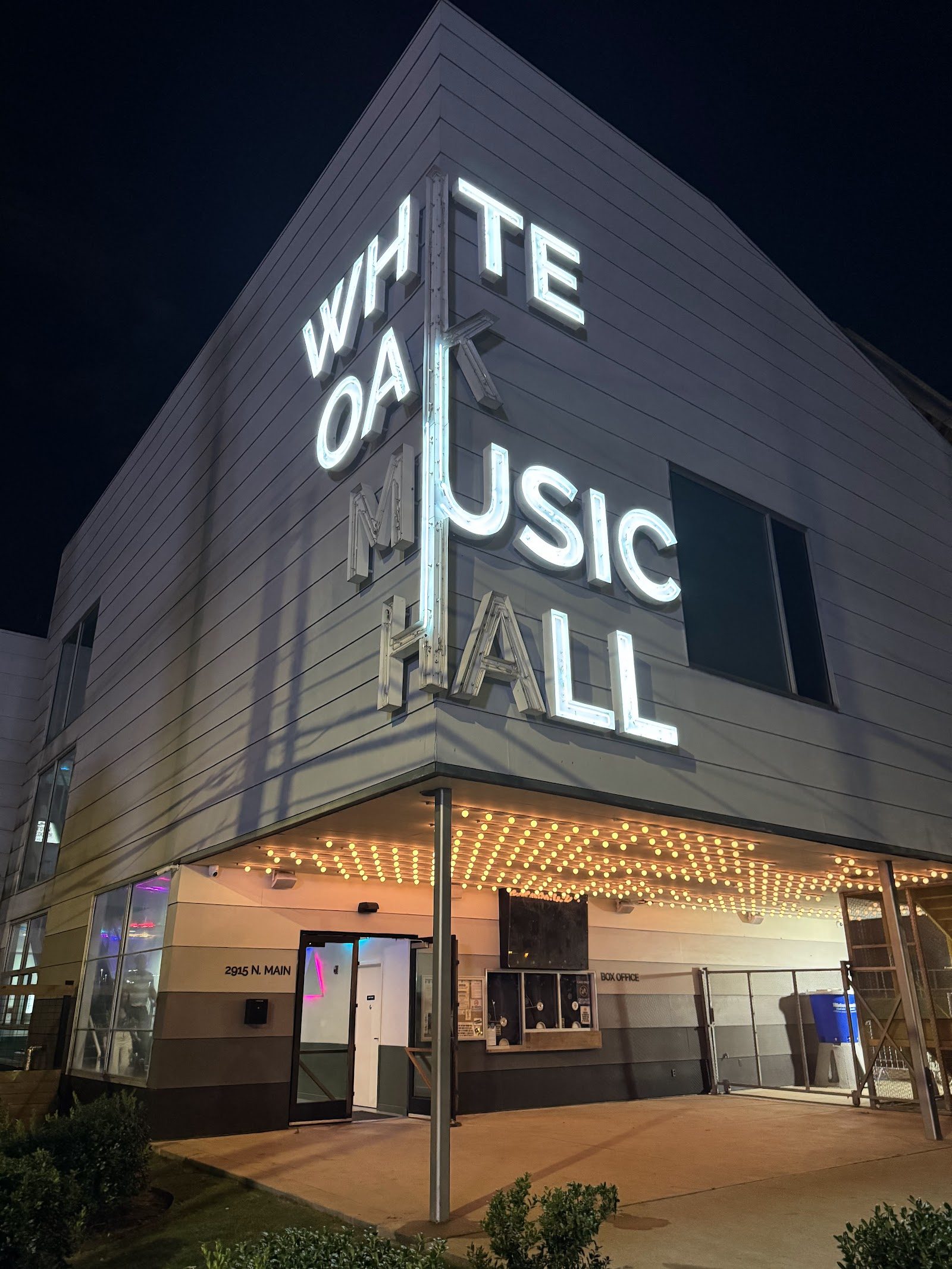 The White Oak Music Hall building at night, its name in tall glowing white letters on the corner above a box office lit by rows of marquee bulbs, with 2915 N. Main painted by the door