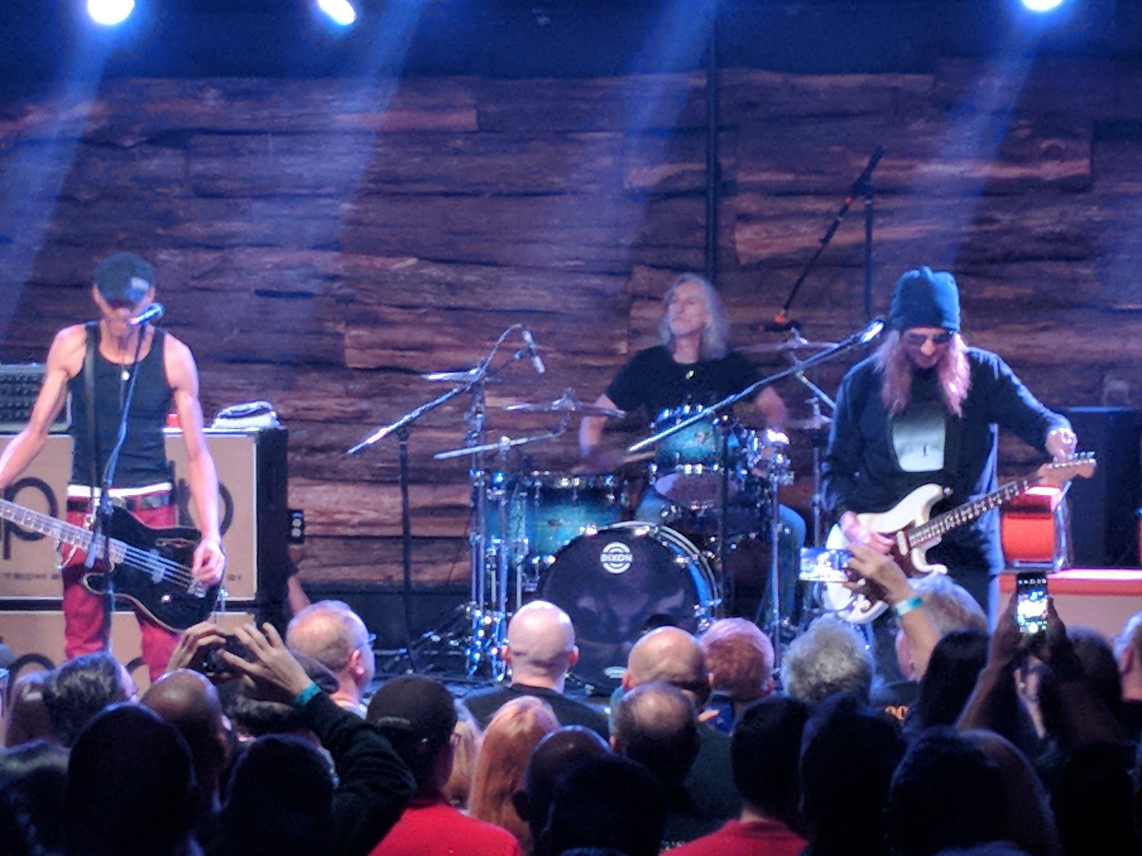 A three-piece band playing under blue stage lights against a rough wooden back wall at Come and Take It Live, with fans holding up phones in front