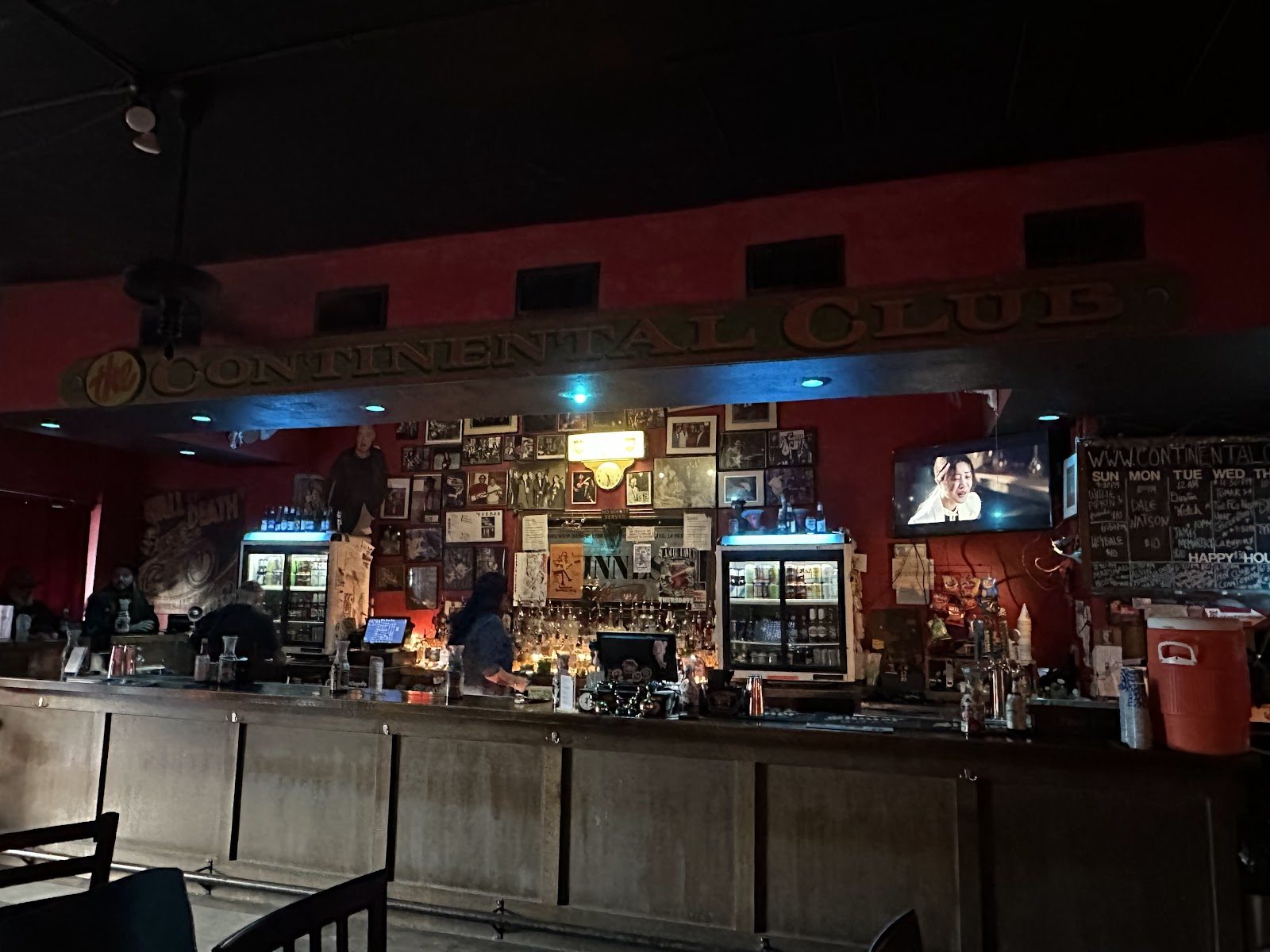 The long wooden bar inside the Continental Club under a carved Continental Club sign, with red walls covered in framed photos and a chalkboard show calendar