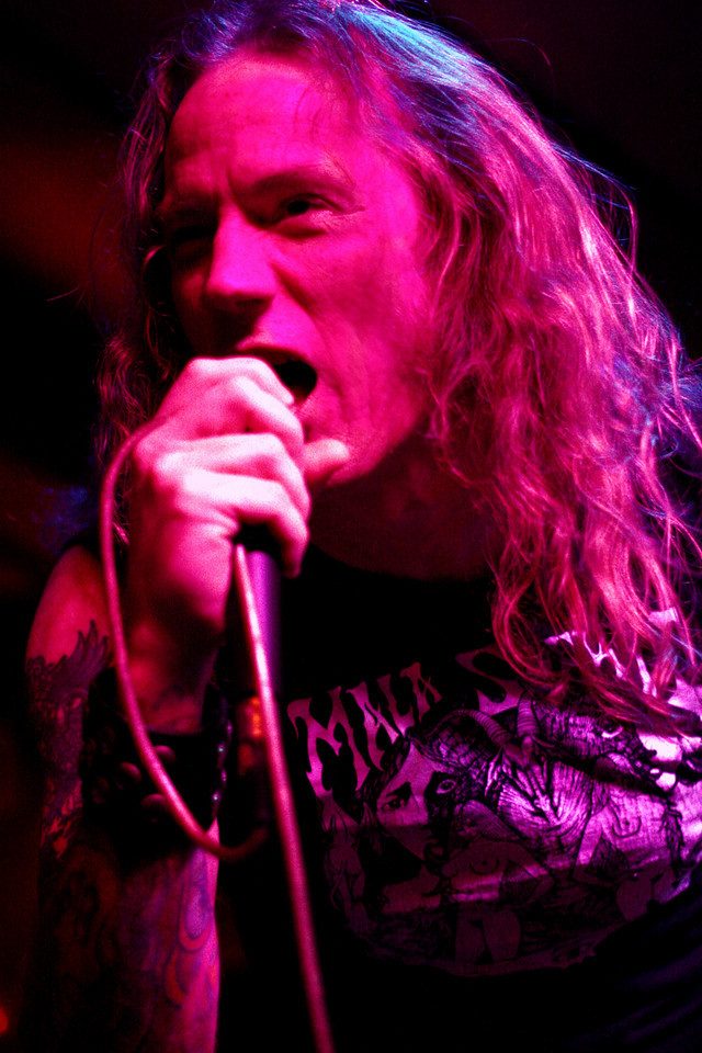 Jason McMaster singing into a handheld microphone under magenta stage light, long hair falling over a black graphic T-shirt